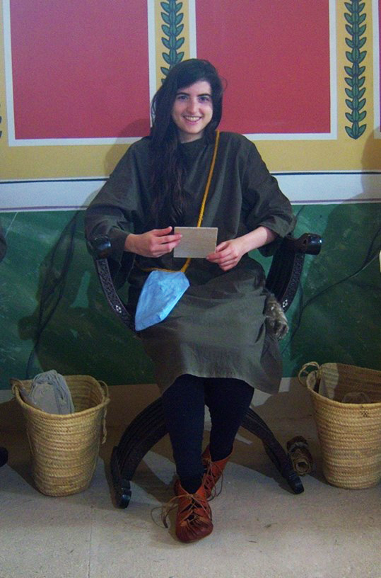 A woman wearing a tunic sitting in a chair in a recreated Roman classroom